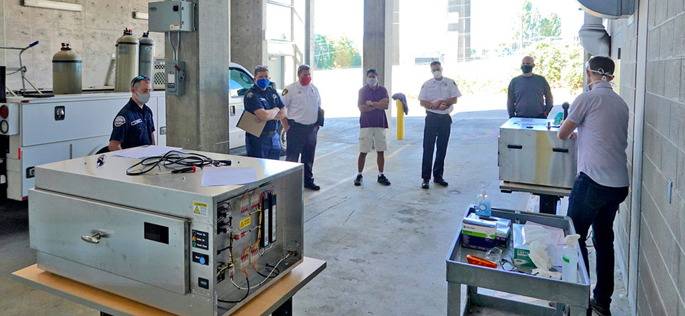A training involving 6 people in masks standing in a distanced circle in a loading dock while UW PhD student Ben Sullivan gestures to a UVC decontamination box.