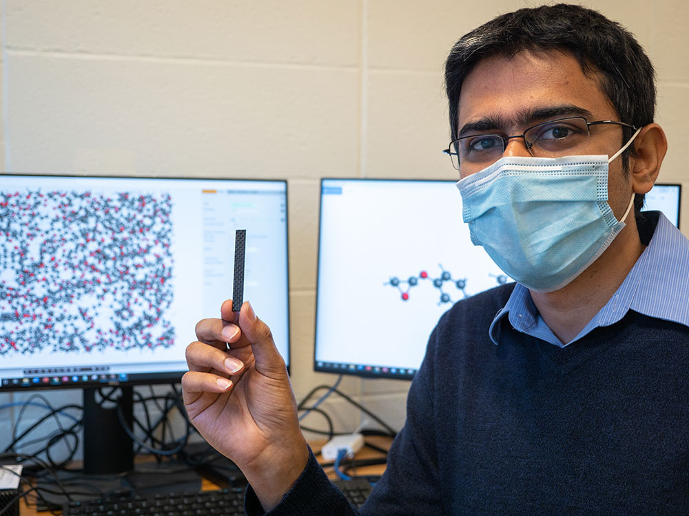 Aniruddh Vashisth holding a sample of a healable carbon fiber composite with computer monitors in the background
