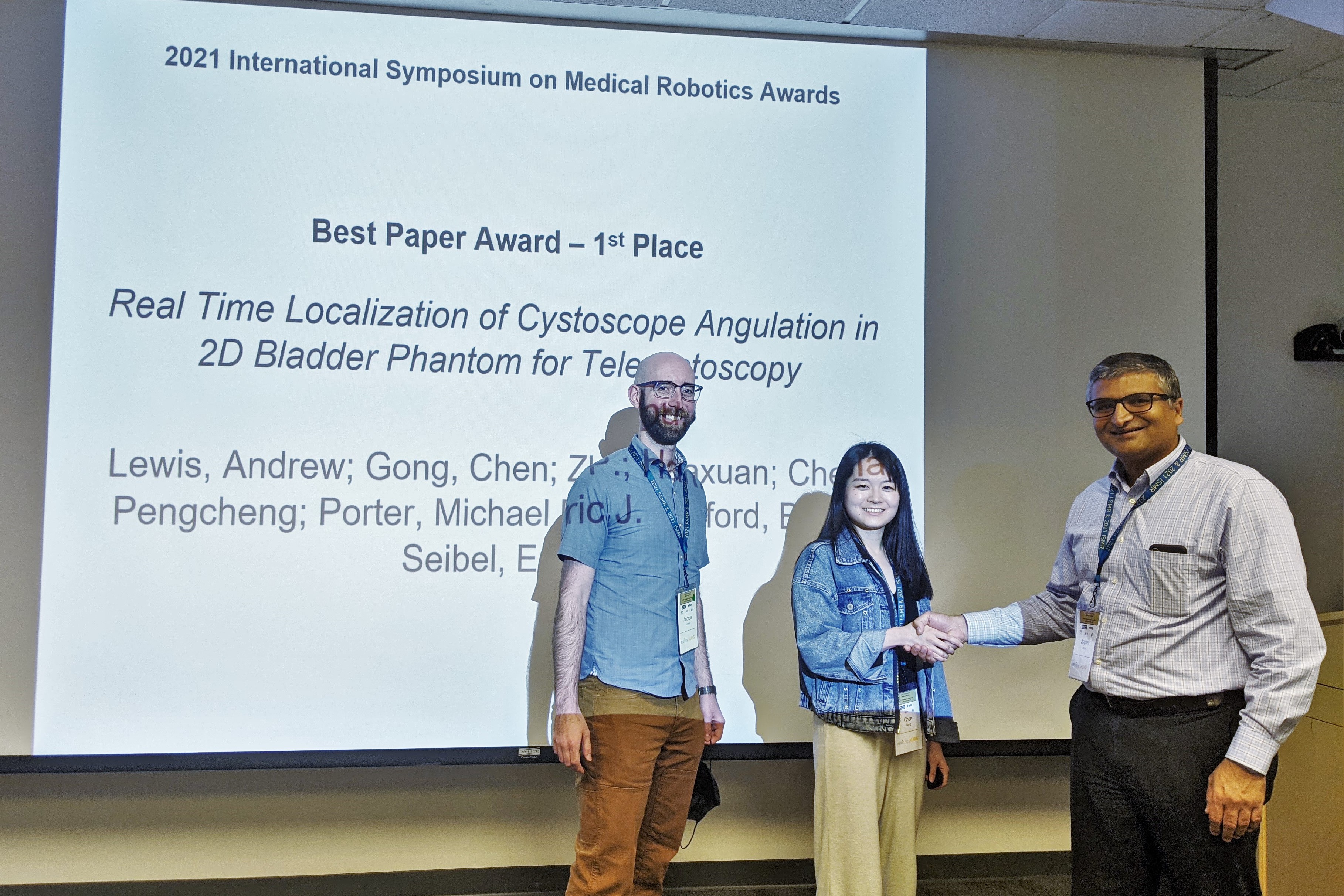 Two people, a man and a woman, both roughly in their 20s, pose for a photo in front of a projector screen that says Best Paper Award - 1st Place. The woman shakes the hand of another man, older than them, to the side of her.