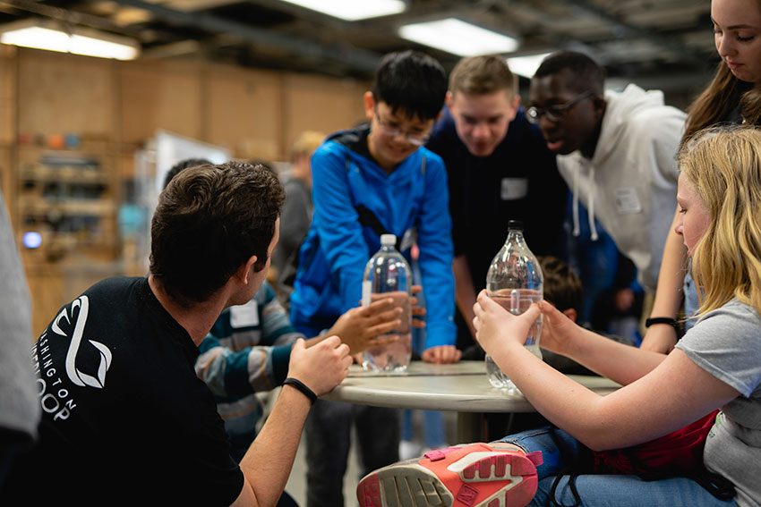 A group of students surrounding a round table while two of them are holding soda bottles. Buffalo is crouching next to them.