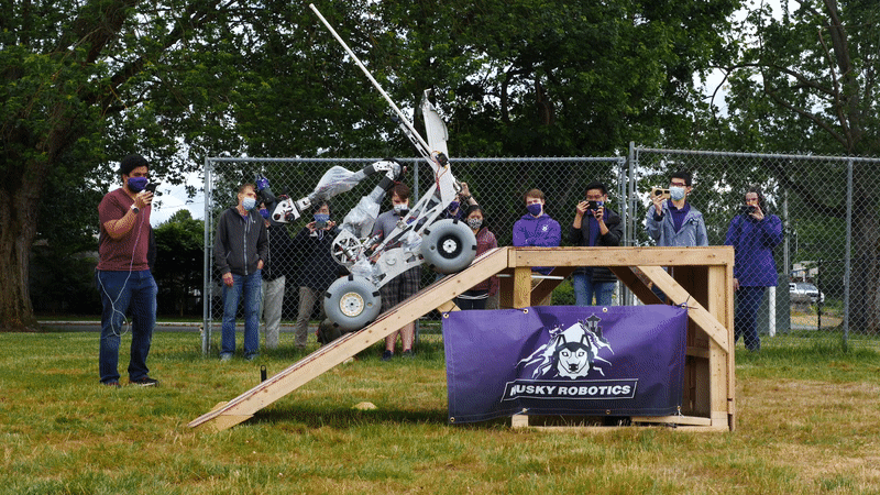 10-second animated image showing a mars rover-like robot rolling slowly up a wooden ramp while onlookers watch