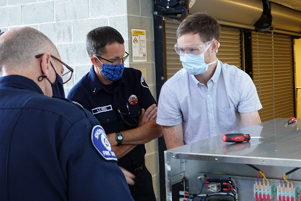 Ben Sullivan with his hands on a box showing it to two fire officials in uniform.