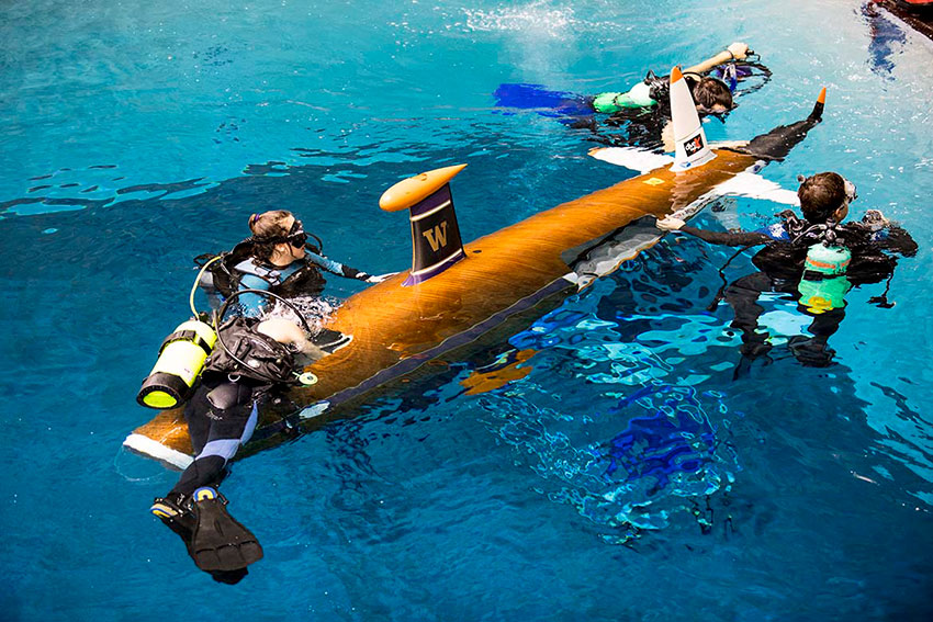 Students in diving suite swimming around a submarine in a pool