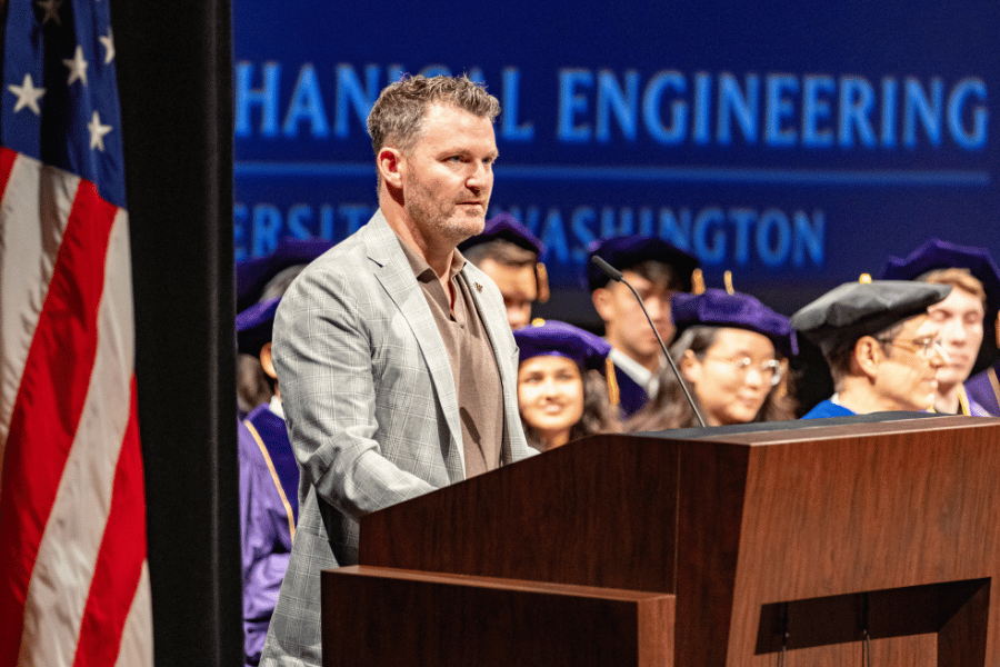 Justin Brynestad stands behind a podium and speaks into a microphone. To his left is part of an American flag and in the background is a group of faculty and Ph.D. students.