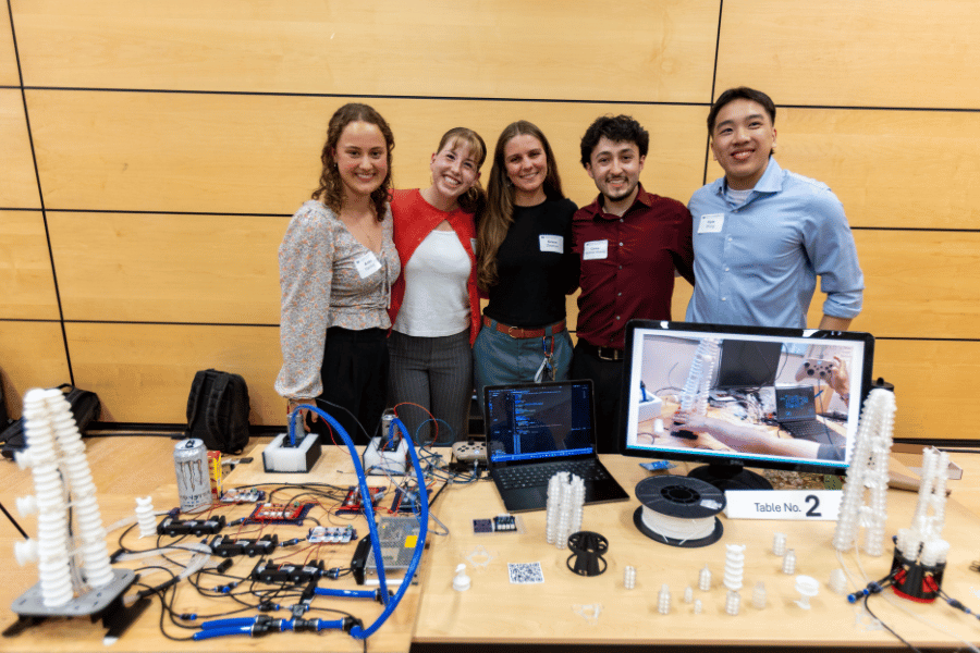 Five students stand behind a table displaying soft, flexible robotics, both individual pieces and put together to resemble an octopus arm.