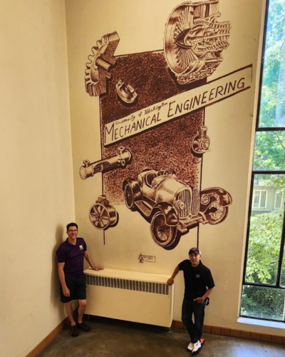 Two people stand in front of a mural in a stairwell.