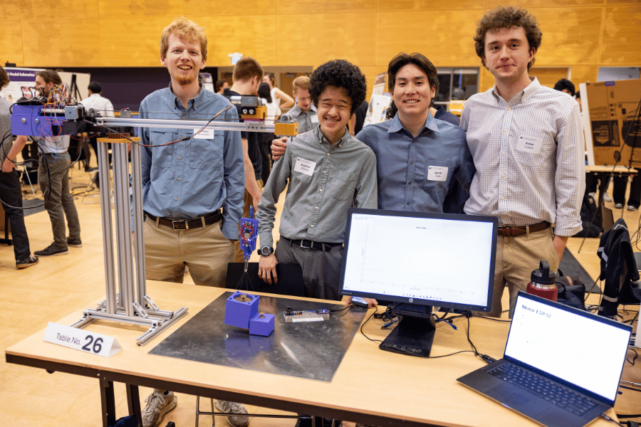Four students stand behind their table at the capstone expo. The table holds a model tower crane that can pick up objects.