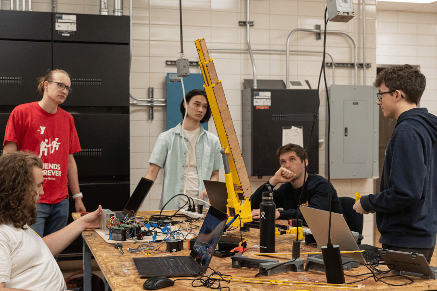 Five students around a table that has model crane parts, a laptop, and wiring on it.