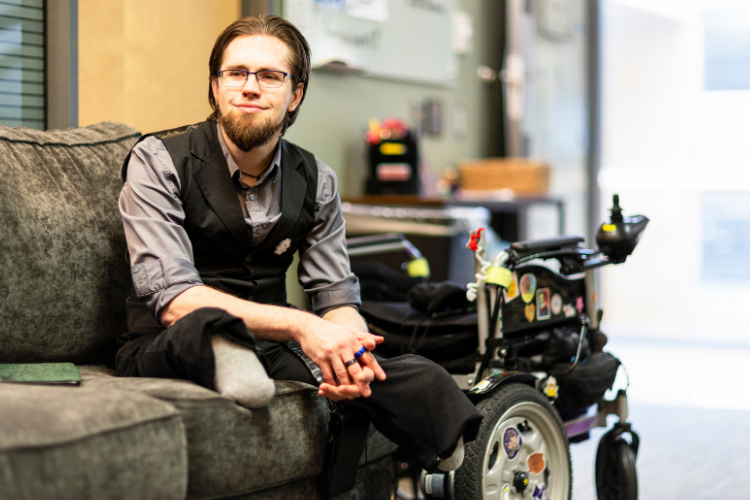 Thomas Brown sits on a couch smiling. He is wearing a vest and dress shirt, and has two amputated legs. His wheelchair is behind him.
