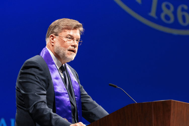Greg Hyslop stands behind a podium and speaks into a microphone.