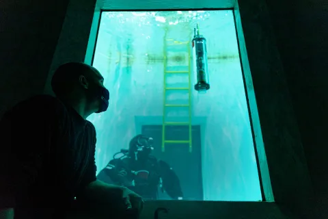 Trevor Harrison looking through the glass pane of a big water tank with a diver and a µFloat inside