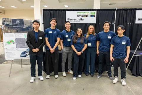 A group of seven individuals stands before a display labeled "Elementrailer" at an indoor event