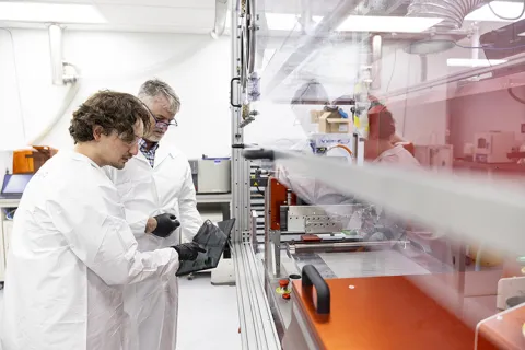 Two people in lab coats and gloves examining a tablet in a laboratory setting, surrounded by scientific equipment.