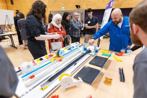 People examining a table with mechanical prototypes and a laptop at an exhibition