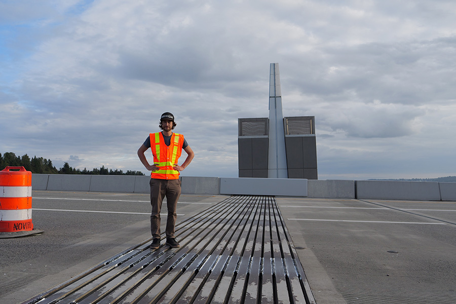 Ph.D. student Sawyer Thomas is wearing a bridge orange safety vest while standing on the SR 520 bridge next to the installed chevron supports.