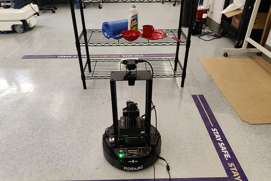 A mobile robot faces a shelf with a pitcher, bowl, plate, cleaning solution, mug and a spoon.