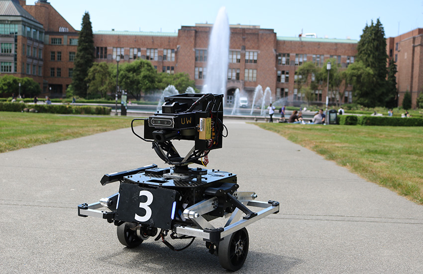 A robot is shown on the sidewalk on the UW campus in front of a fountain.