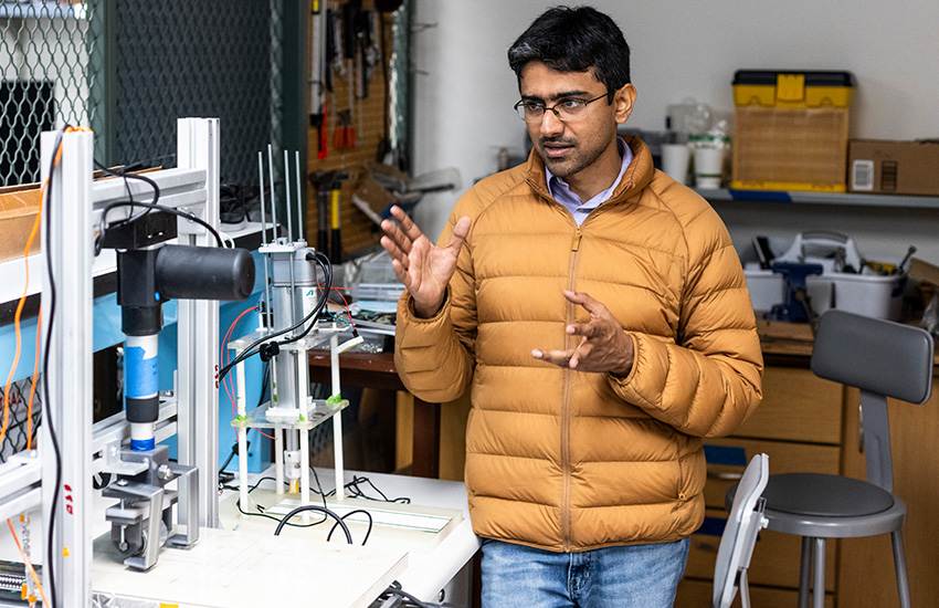 Assistant Professor Aniruddh Vashisth stands in a lab environment next to two machines used for automated welding