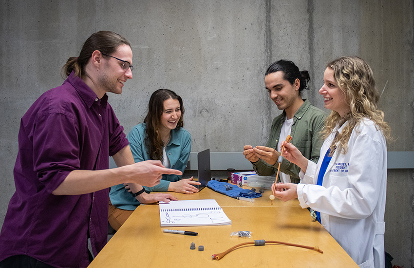 Four CathConnect team members gather around a table. Two are holding parts of the CathConnect device, one is smiling on a laptop, and another person is pointing at the device.