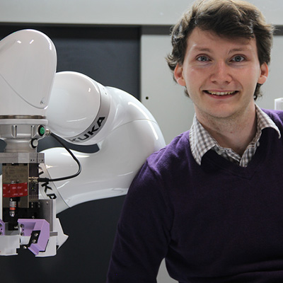 Ian Good smiles and stands to the right of a robotic gripper