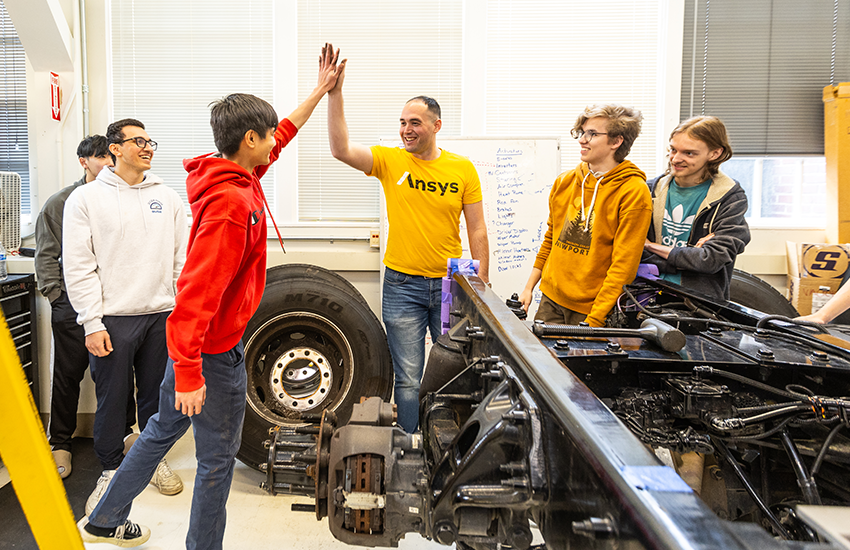 Six members of the E-Truck team stand near the back of the truck. The wheel has been disassembled. Two people are high-fiving.