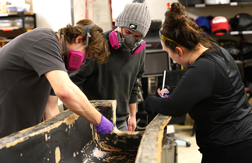 Two students wearing large masks are sanding and examining a large structure that will be part of the Formula car. A third student is taking notes.