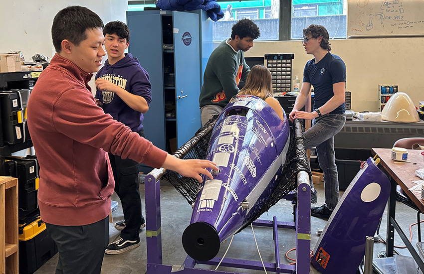 Four students stand near a purple human-powered submarine that they are working on. One student is partially inside the submarine.