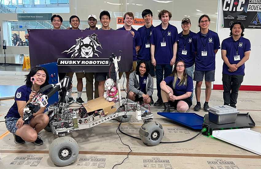 The Husky Robotics team wearing matching shirts, holding up a sign, and posing with their robot.