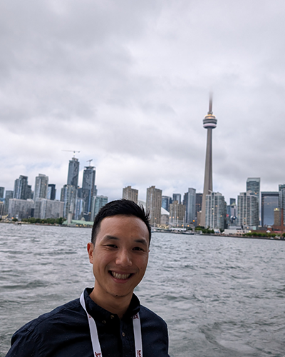 A man stands smiling against a backdrop of water and a city skyline.