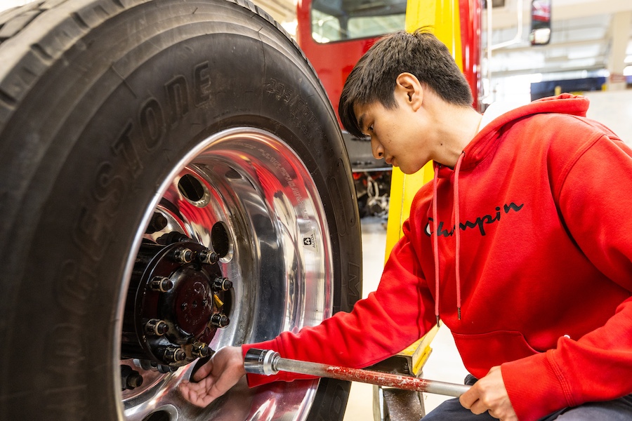 A student holds a tool near a wheel of the truck to begin disassembling it.