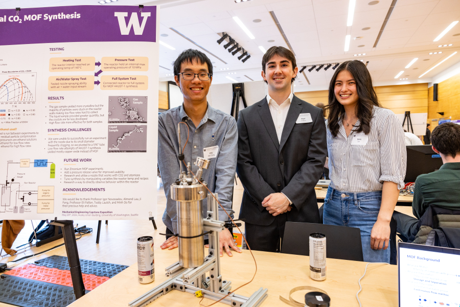 Three students stand behind a vessel that is a reactor for synthesizing materials.