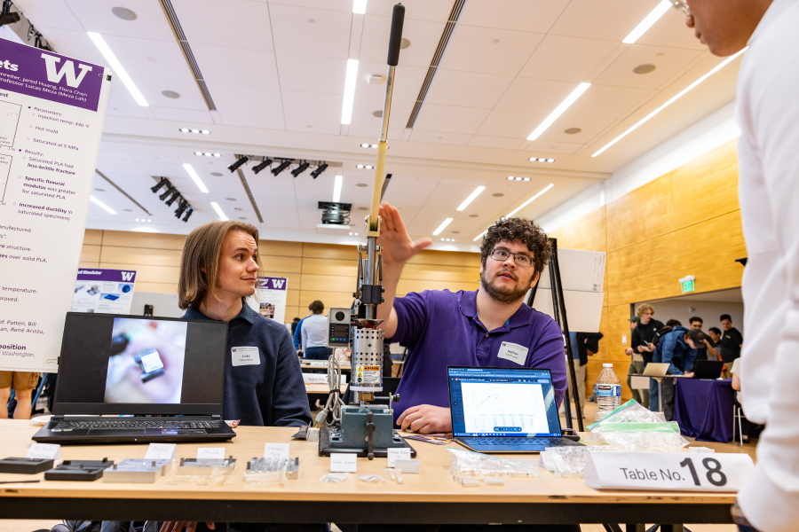 Two students sit behind a table that showcases small plastic pieces and an injection molding machine.