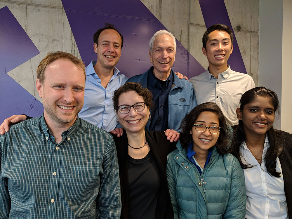 Group photo of seven people standing in two rows against a wall with a W sign