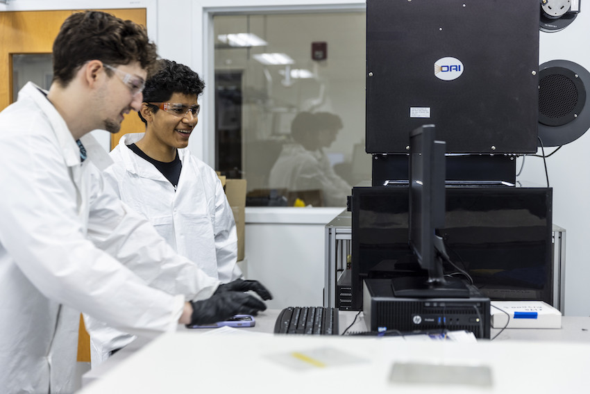 Two men in white coats and goggles looking at a computer