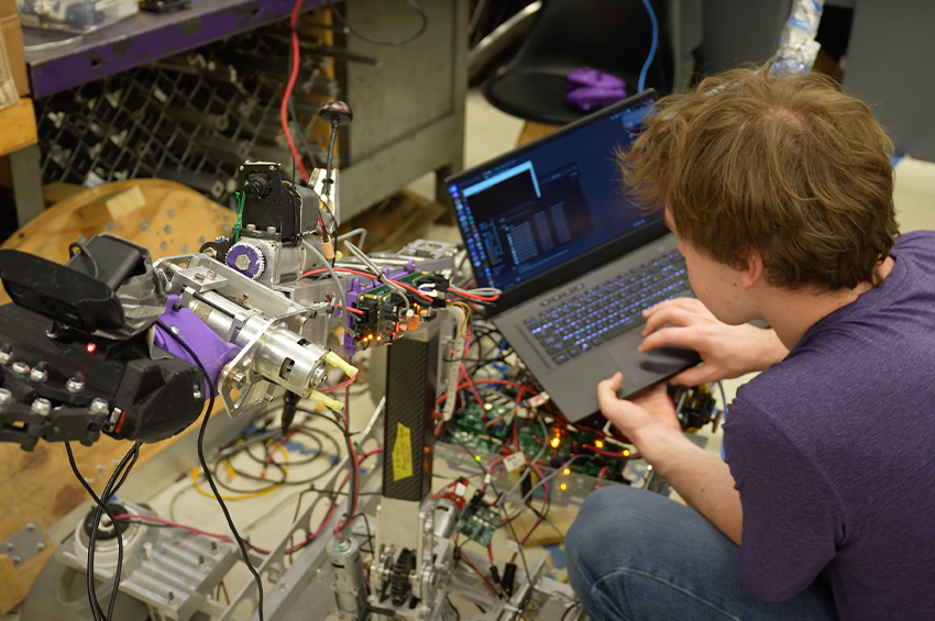 A person in a purple shirt works on a laptop connected to a complex robotic device with wires and mechanical parts in a workshop.
