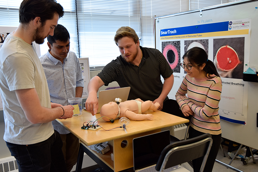 Four individuals work on a project involving a medical dummy and electronic components on a table.