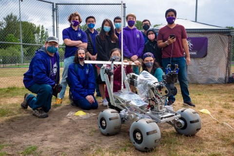 A team of 12 people in covid masks stand outdoors in a group behind a robot that looks like a mars rover