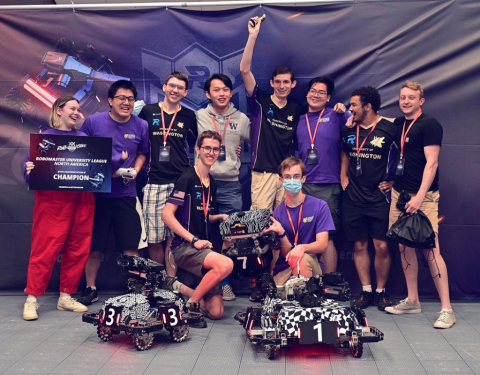 Ten students, several wearing purple, stand in a group with two small wheeled robots in front of them