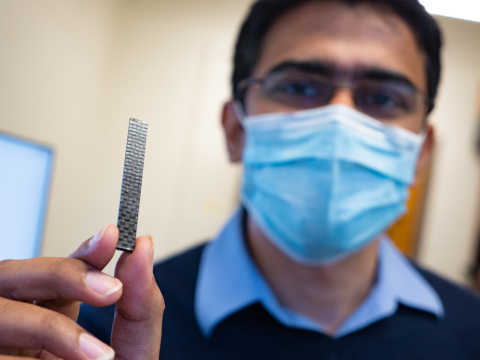A man with dark skin holds a rectangular black chip of carbon fiber material in the foreground and his masked face is out of focus in the background