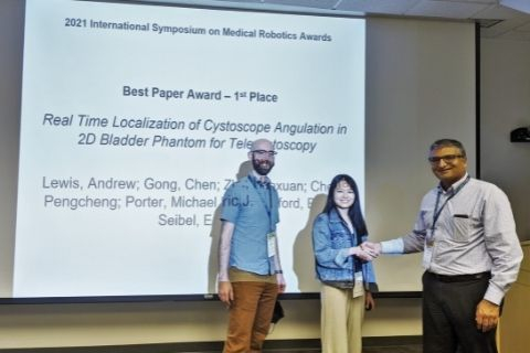 Two people, a man and a woman, both roughly in their 20s, pose for a photo in front of a projector screen that says "Best Paper Award - 1st Place". The woman shakes the hand of another man, older than them, to the side of her.