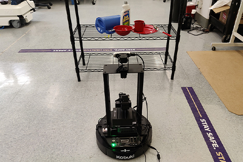 A mobile robot faces a shelf with a pitcher, bowl, plate, cleaning solution, mug and a spoon.