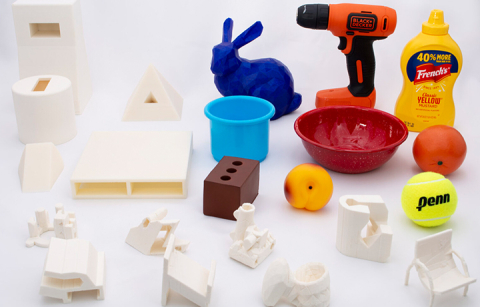 22 objects on a table top. Objects include white 3D printed shapes and also random household items such as a drill, a mustard container, a bowl and a tennis ball