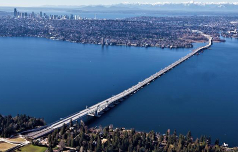 Aerial view, looking west, of the new SR 520 floating bridge, with the Medina shoreline in the foreground and downtown Seattle skyline in the distance.